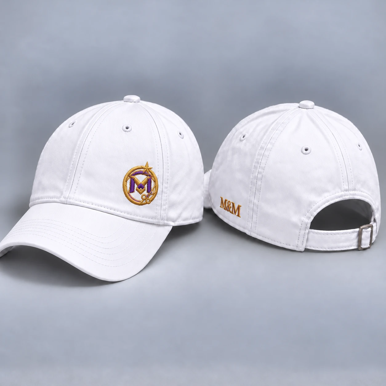 White baseball cap with a compact gold-and-purple emblem embroidered off-center above the brim, M&M lettering low on the side above the bottom edge, and a plain back